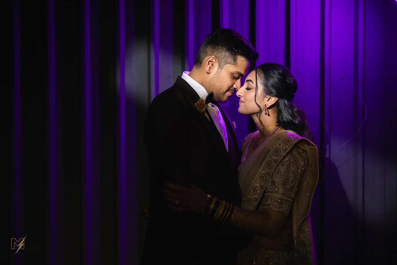 Groom in a black suit embracing bride in a gold embroidered saree against a purple backdrop under spotlight.