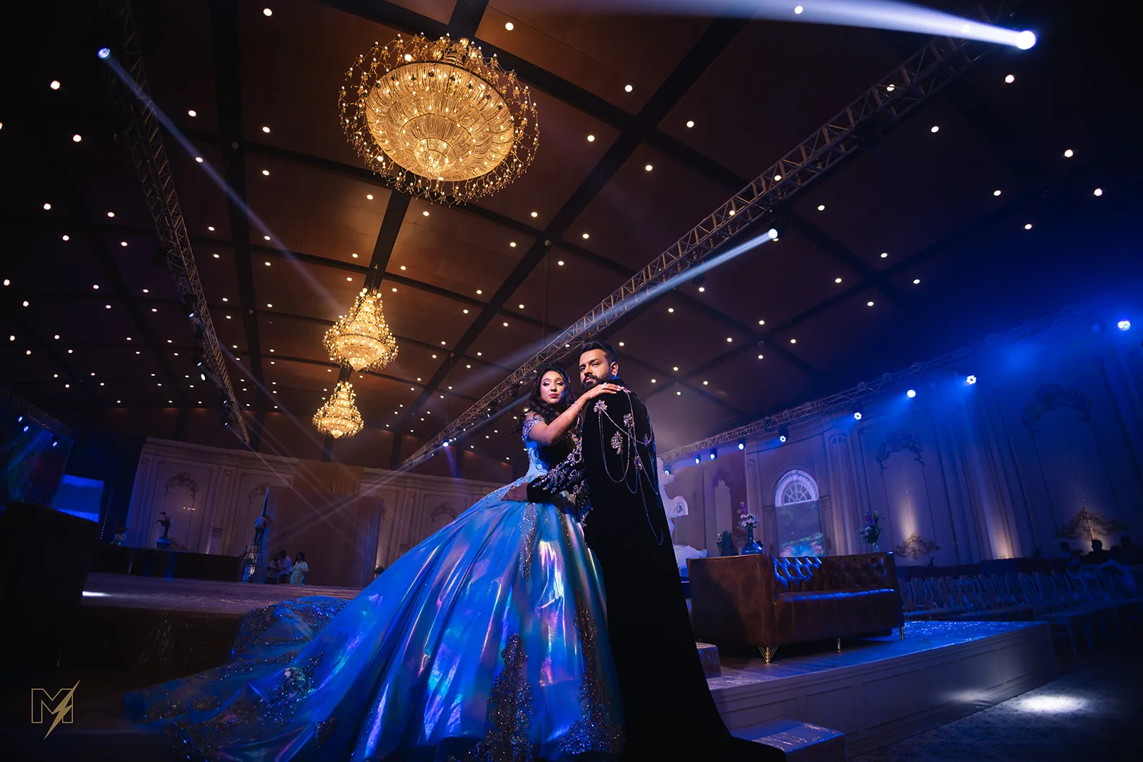 Bride in a voluminous shimmering blue ballgown and groom in a embellished black sherwani posing on stage under luxury chandeliers.