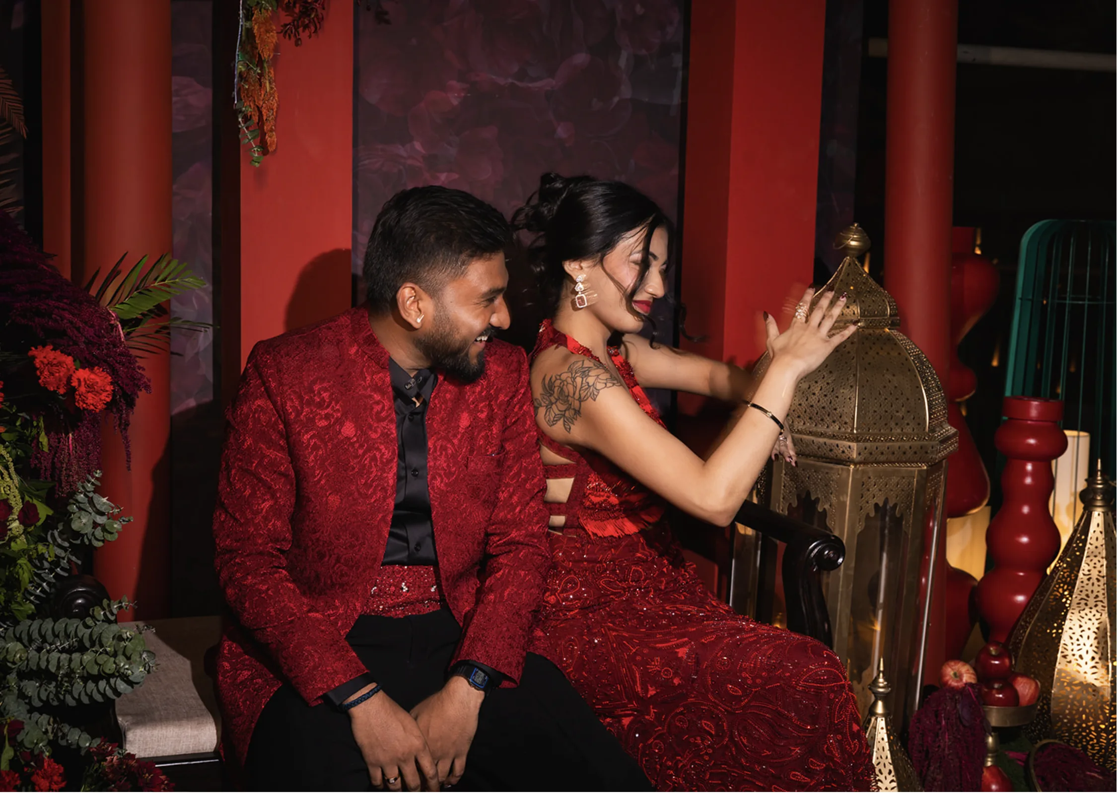 A bride and groom wearing color-coordinated deep red outfits sharing a playful candid moment next to ornate brass lanterns.
