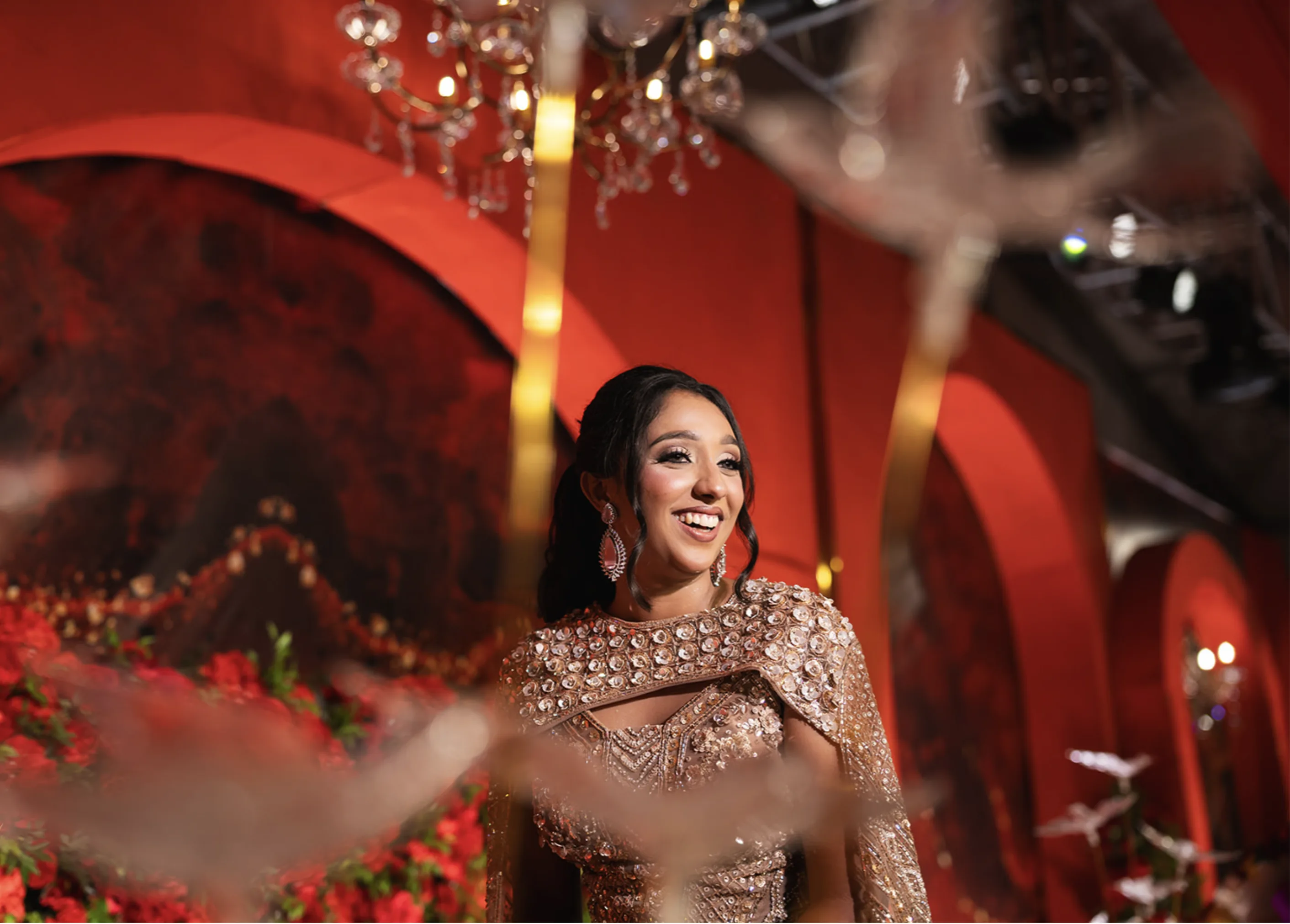 Smiling Indian bride in a heavily embellished champagne gold outfit with a cape against a red arch background with chandeliers.